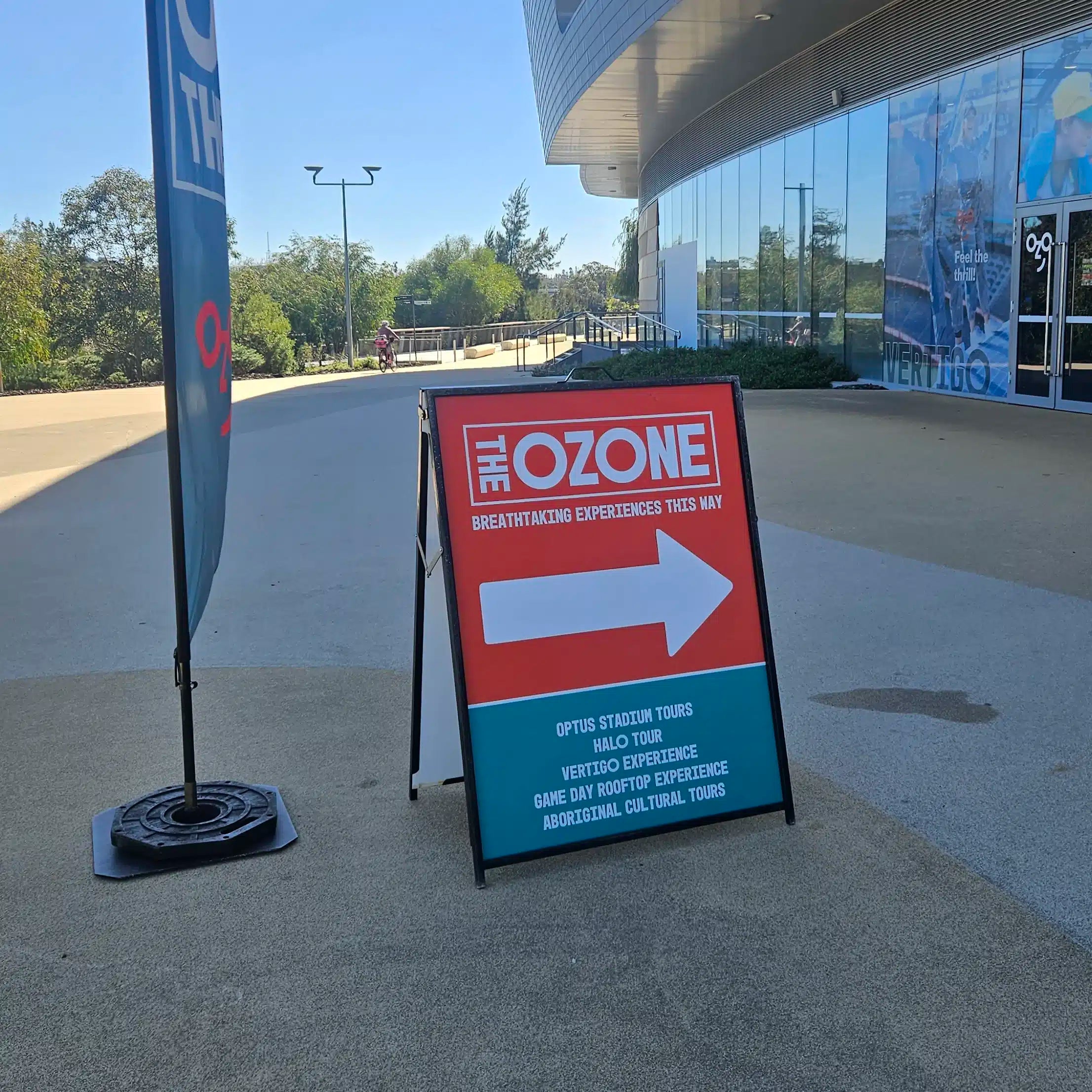 Black A-frame sign outside Optus Stadium directing visitors to stadium tours, beside a teardrop flag