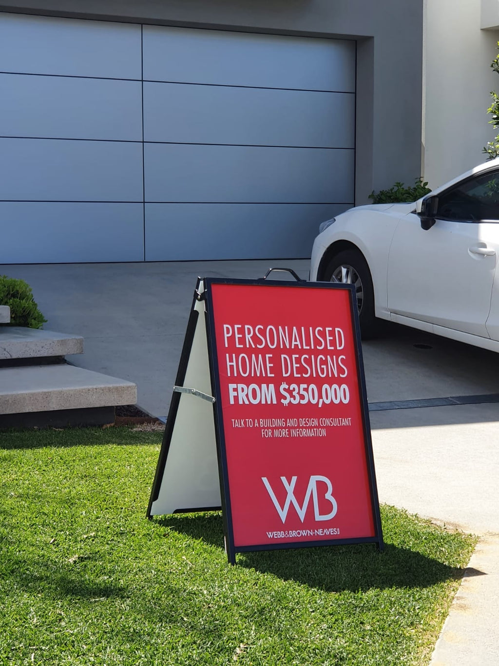 Red corflute A-frame sign on a lawn outside a display home, advertising personalised home designs
