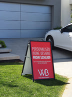 Red corflute A-frame sign on a lawn outside a display home, advertising personalised home designs