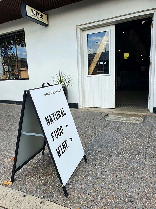 Black A-frame on a footpath outside a restaurant reading Natural Food and Wine with an arrow