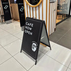 Black A-frame in a shopping centre walkway reading Cafe Now Open