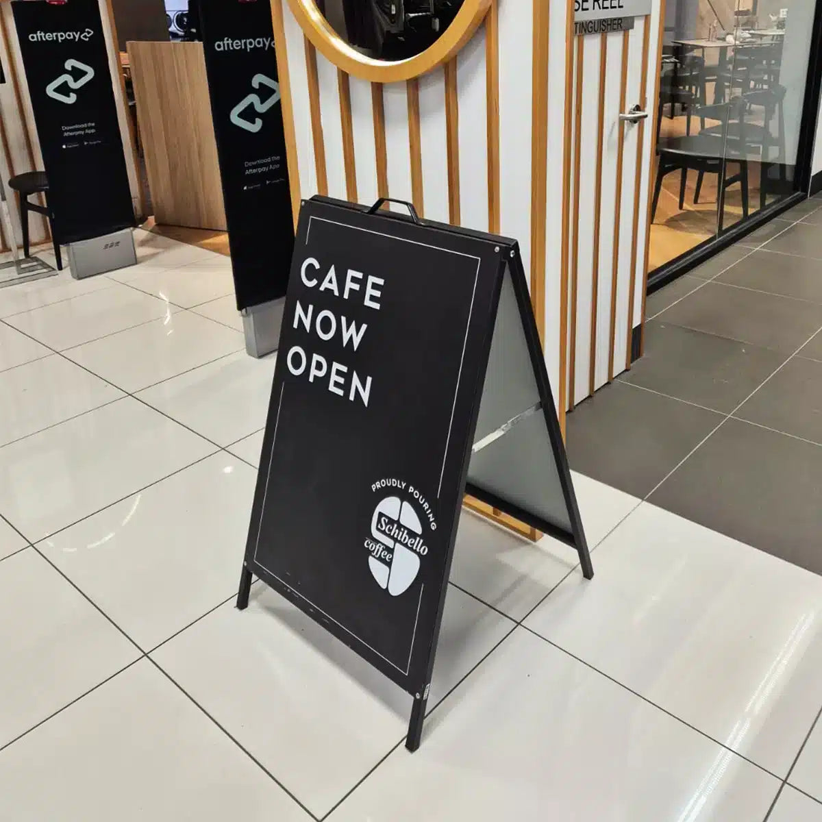Black A-frame in a shopping centre walkway reading Cafe Now Open