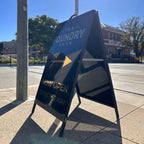 Black A-frame on a footpath with a Now Open arrow sign pointing to a coffee shop