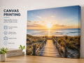 A canvas wall print showing a beach boardwalk leading to the ocean at sunset, displayed on a tabletop next to a white wall with text reading 'CANVAS PRINTING' and small decorative plants and books.