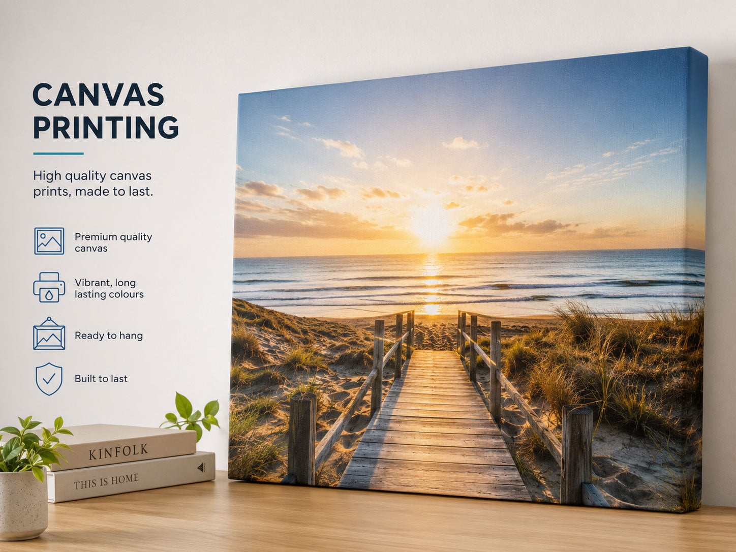 A canvas wall print showing a beach boardwalk leading to the ocean at sunset, displayed on a tabletop next to a white wall with text reading 'CANVAS PRINTING' and small decorative plants and books.