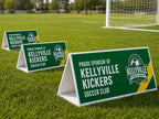 Three green triangle roster signs standing on the grass beside a soccer goal, printed for a club sponsor